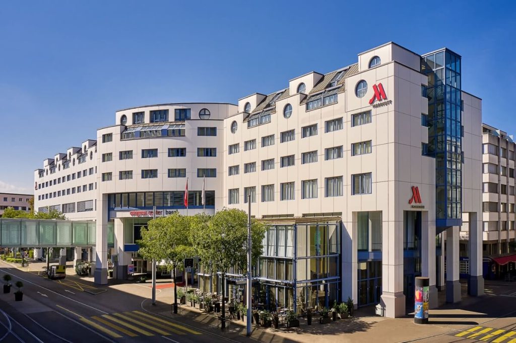 Exterior view of a modern multi-story hotel building with the “Marriott” logo prominently displayed, featuring large windows, a glass elevator, and a skybridge on the left side, set against a clear blue sky.