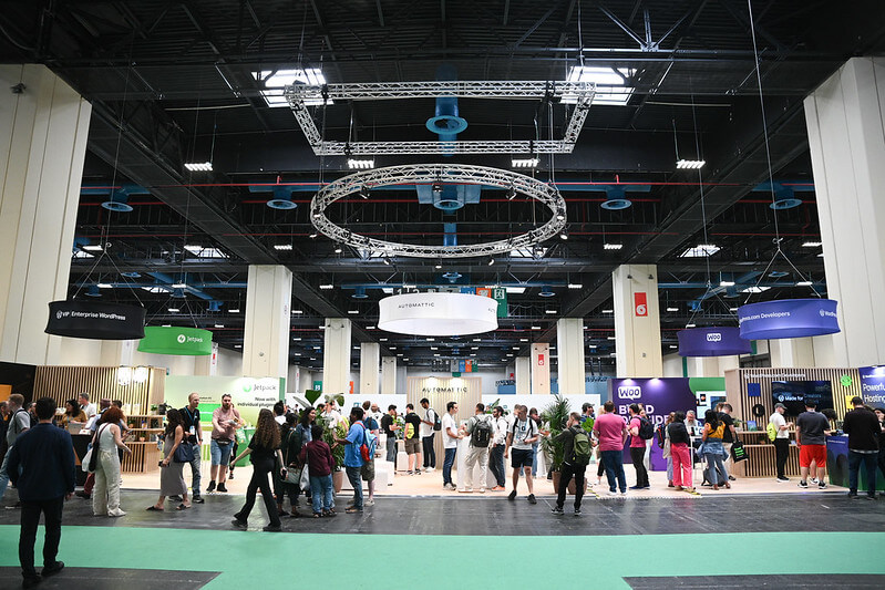 A spacious exhibition hall with multiple booths and attendees gathering and conversing at WordCamp Europe 2024.