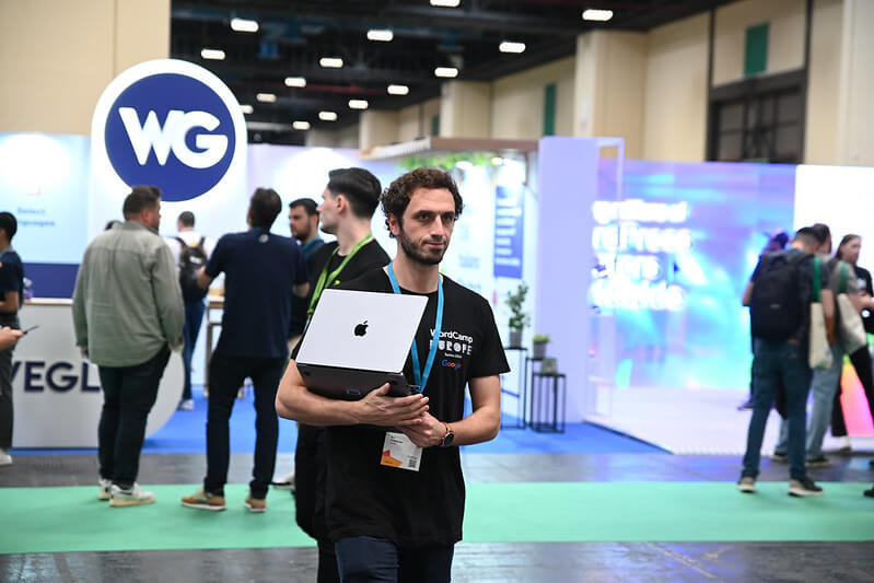 An attendee carrying a laptop, walking through an exhibition area filled with booths and participants at WordCamp Europe 2024.