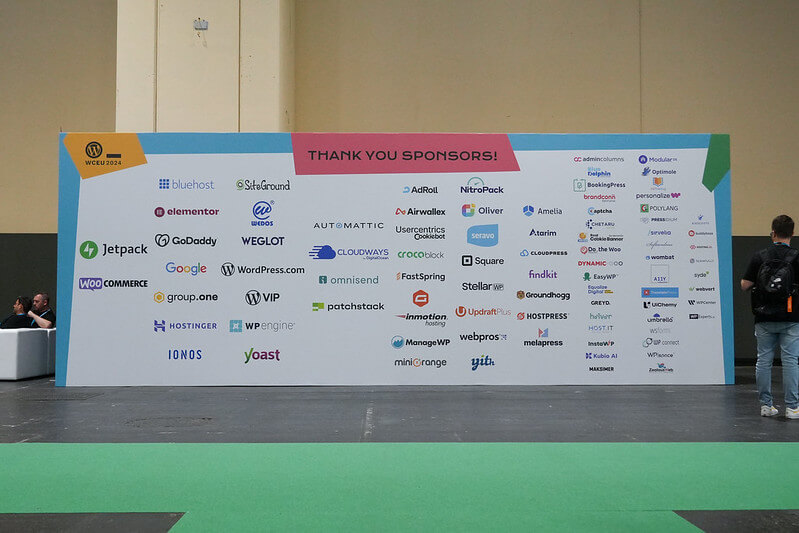 A large display wall featuring the names of sponsors at WordCamp Europe 2024, with attendees passing by.